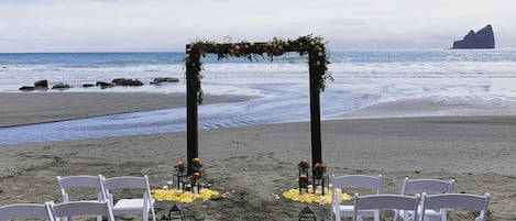 Une cérémonie de mariage organisée sur la plage, avec des chaises blanches et une arche décorée de fleurs.