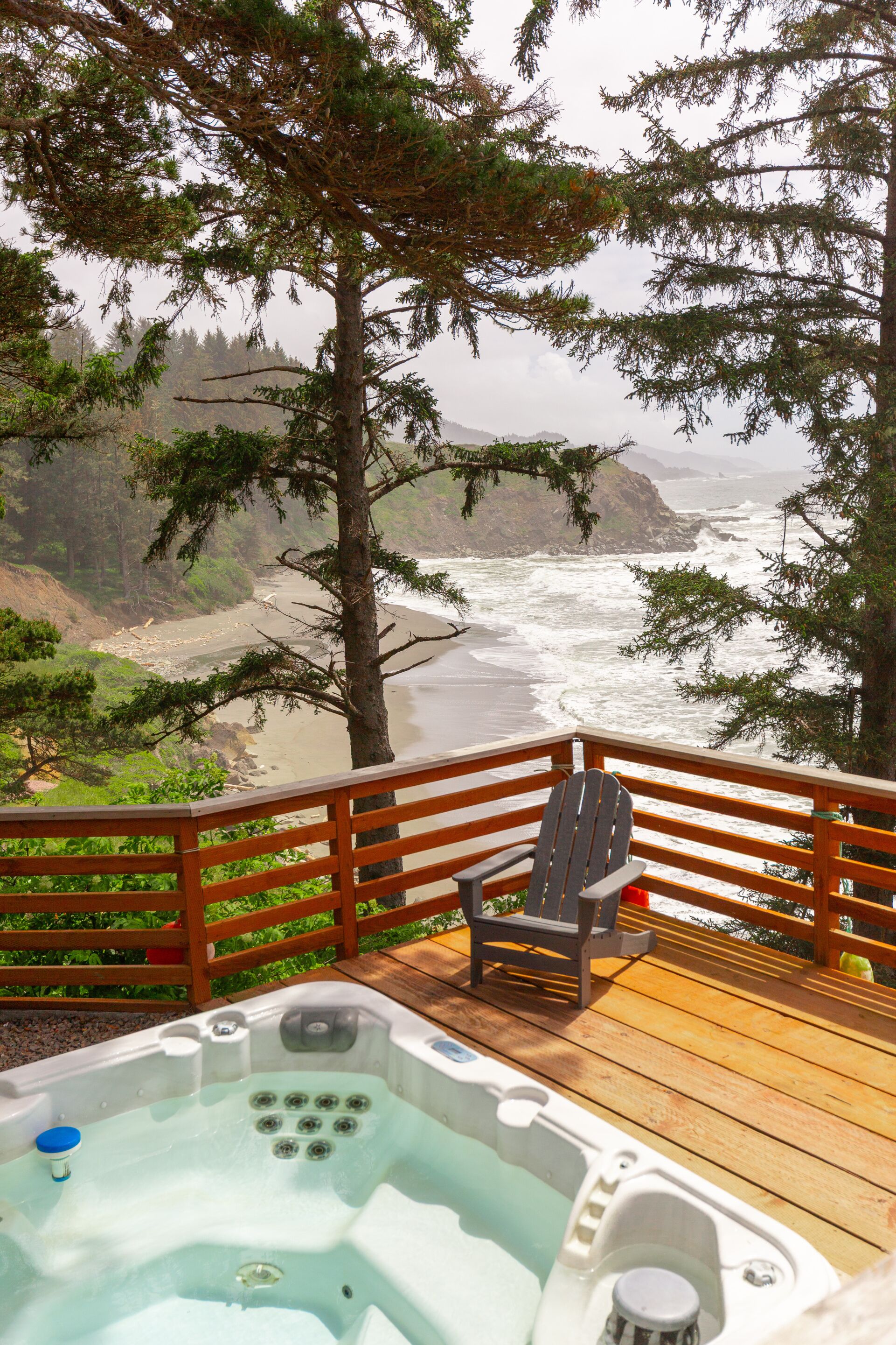 A wooden deck with a hot tub and a chair, overlooking a beach and ocean.