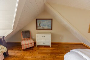 A bedroom with a sloped ceiling, a painting on the wall, a striped chair, a white dresser, and wooden flooring. - Nautilus - Oceanfront Nantucket Style Home (Gold Beach)