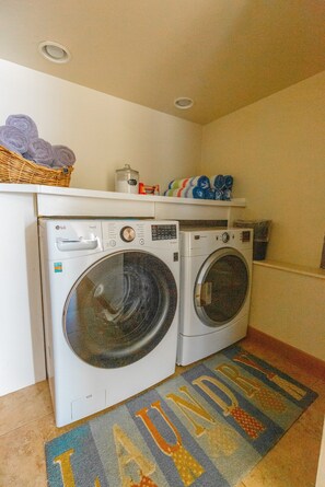 A laundry area with two front-loading washing machines, a shelf with laundry supplies, and a colorful towel mat. - Nautilus - Oceanfront Nantucket Style Home (Gold Beach)