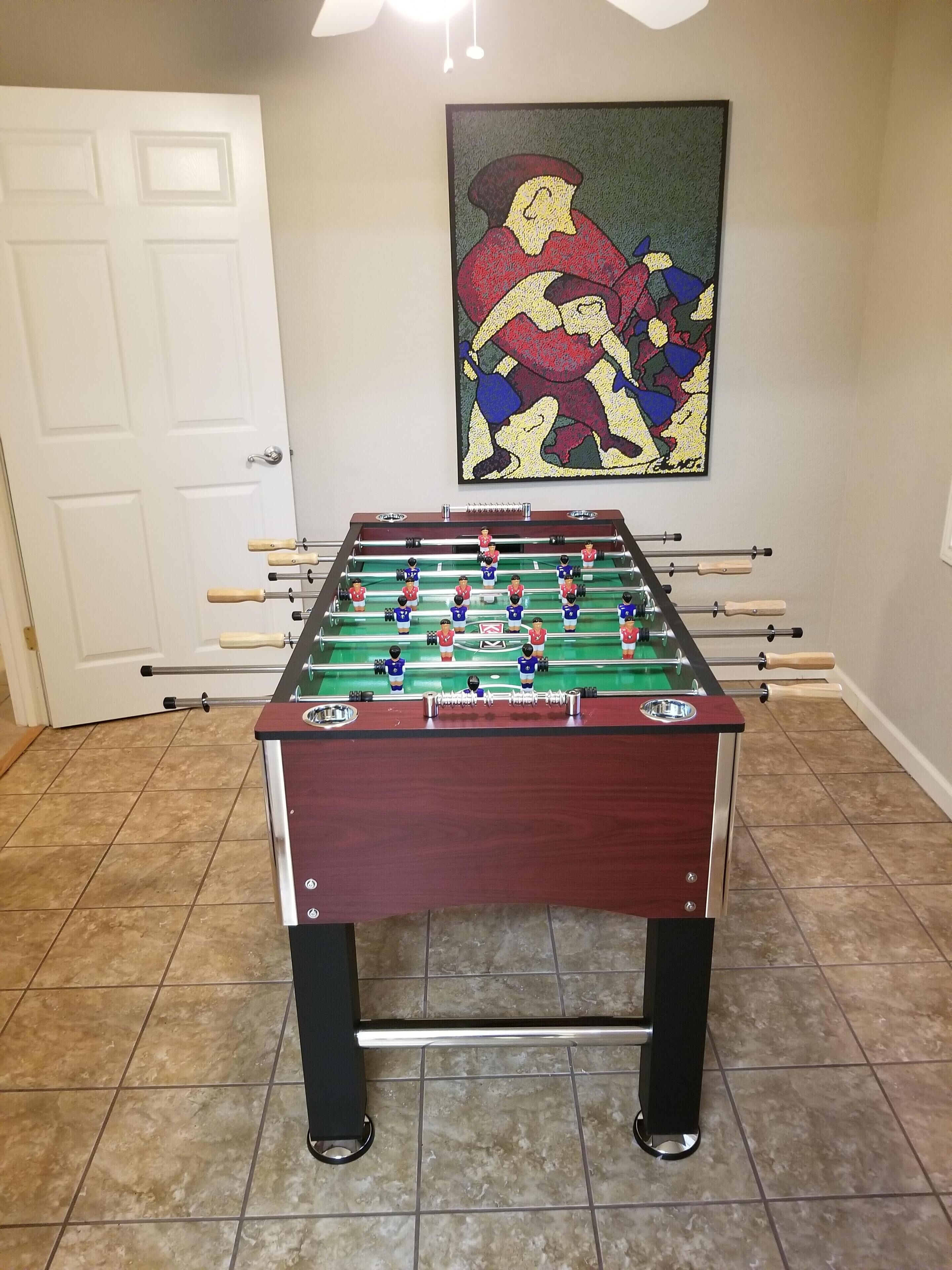 Een tafelvoetbaltafel in een kamer met betegelde vloer, een witte deur en een kleurrijk schilderij aan de muur.