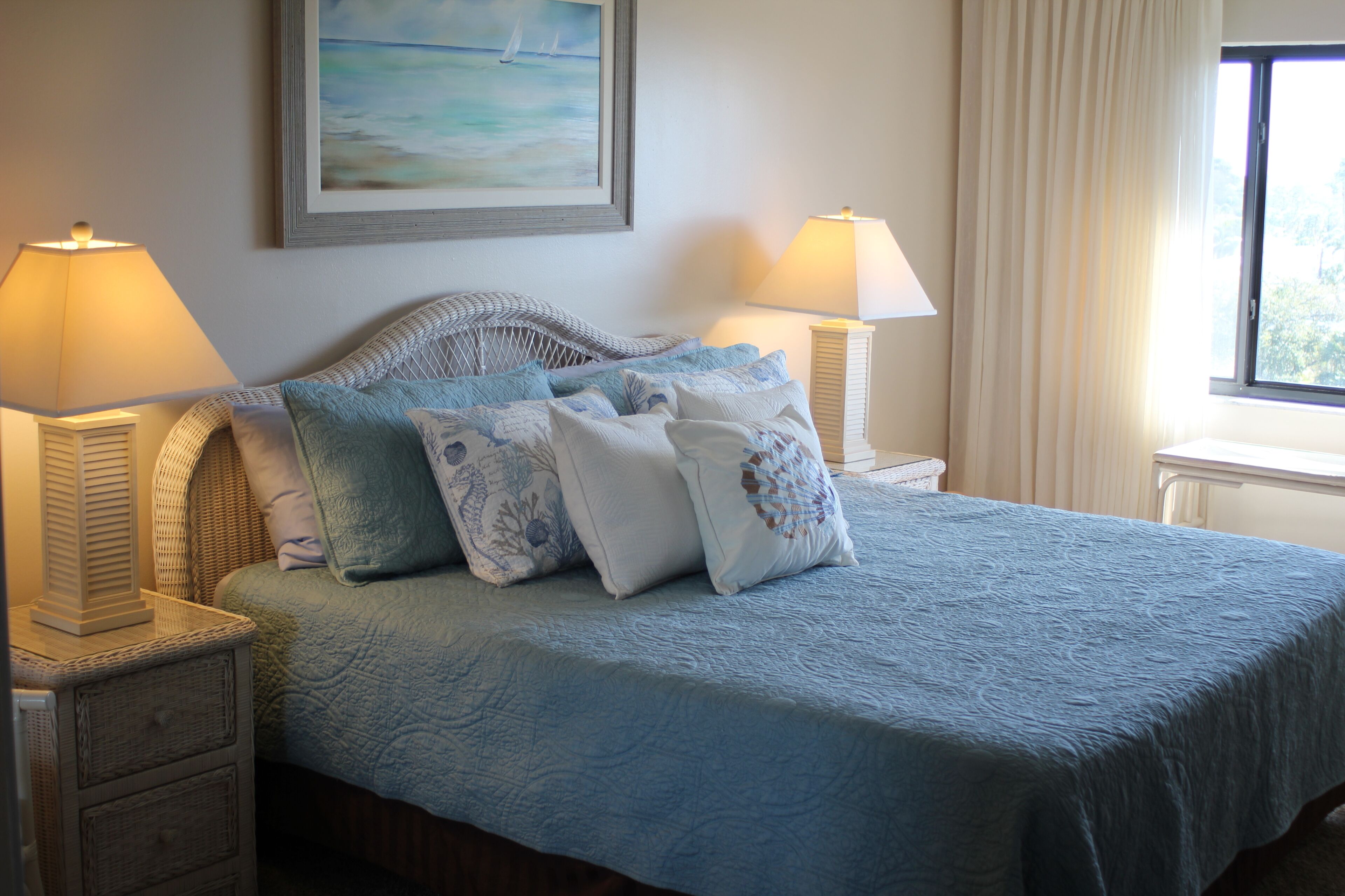 Una cama bien hecha con colcha azul y almohadas decorativas, flanqueada por dos mesitas de noche con lámparas, en una habitación con un cuadro en la pared y una ventana con cortinas.