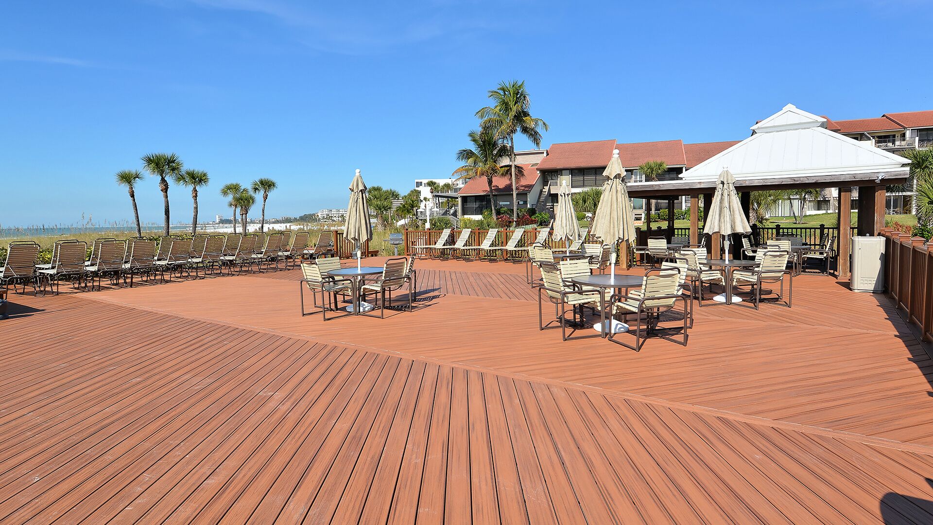 Terraza de madera con mobiliario exterior y sombrillas, con vistas a una playa y un edificio de techo rojo.