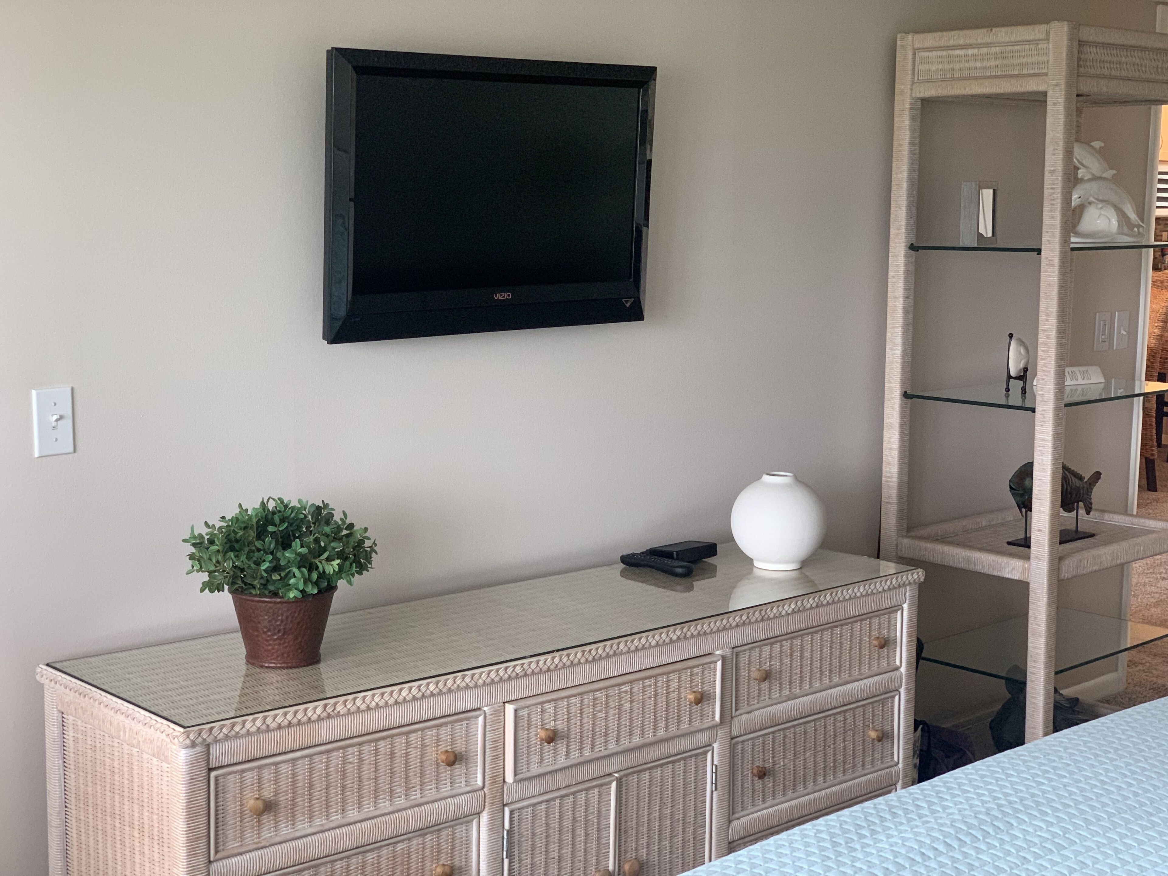 A room with a flat-screen TV mounted on the wall, a wicker dresser with drawers, a small potted plant, and a white vase.