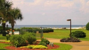 A landscaped garden with a pathway, palm trees, and a view of the ocean. - Low Rates ! Gulf View; Palms, Grnd Flr; Weekly discounts (Gulf Shores)