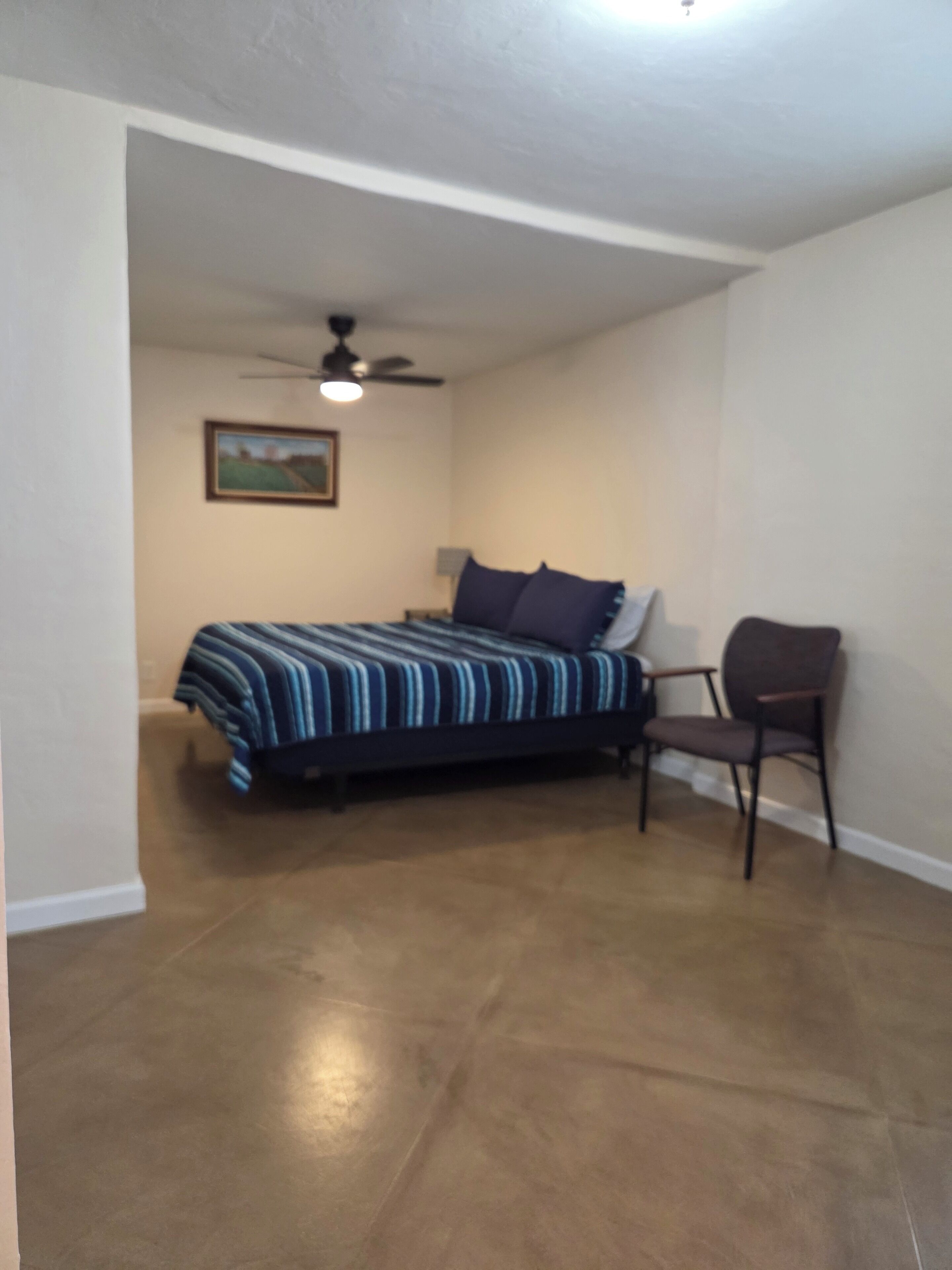 Un dormitorio con una cama, una silla, un ventilador de techo y un cuadro en la pared.