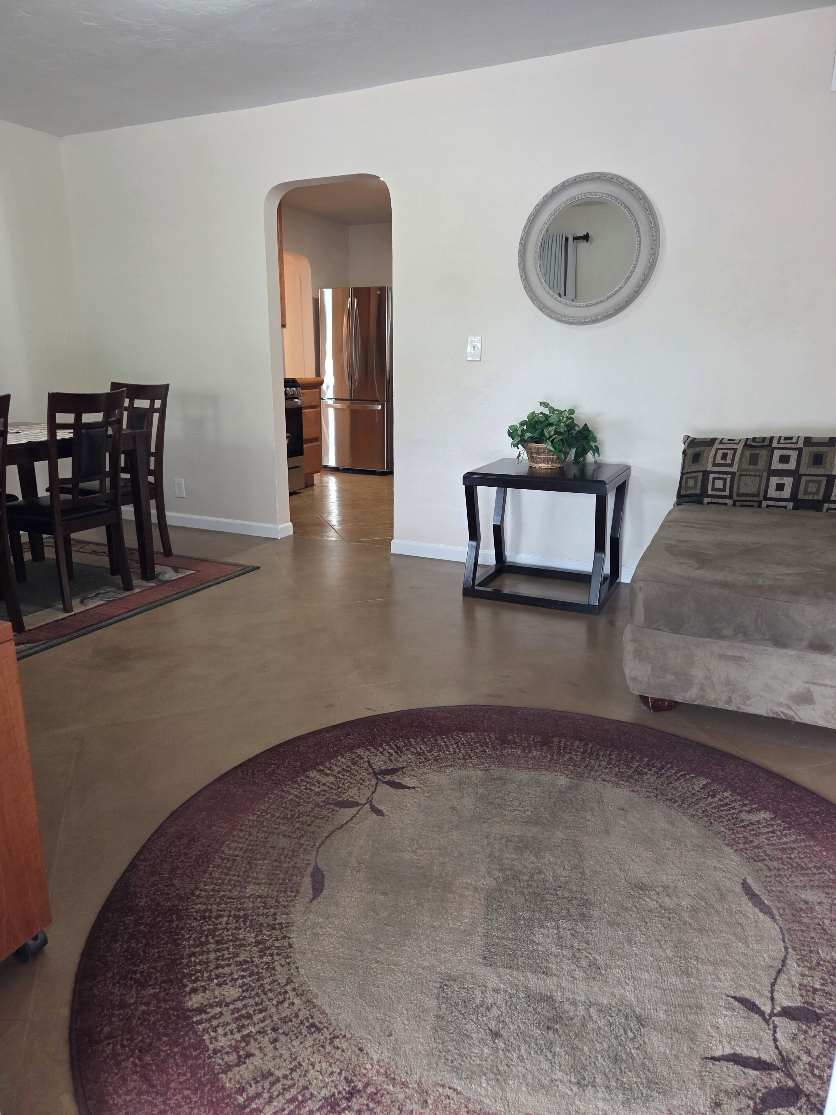 Una sala de estar con comedor, un sofá, una mesa de centro, un tapete, un espejo y una cocina al fondo.