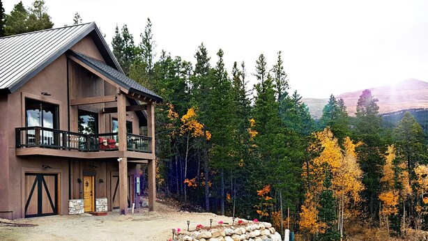 Un chalet à deux étages doté d’un balcon, d’une entrée en pierre et offrant une vue sur une chaîne de montagnes boisées.