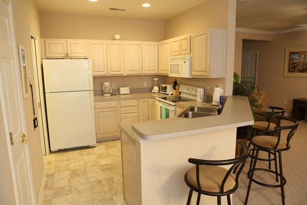A kitchen with a refrigerator, microwave, toaster, and coffee maker. There are three bar stools and a sink. - Relaxing 2 BR/2 BA Condo with 2 Porches & Pool (Rehoboth Beach)