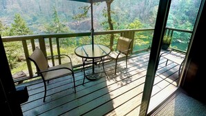 A balcony with a table and chairs, a large umbrella, and a view of a forested area. - River Vista Homes: Perfect for Fishing, Swimming, and Relaxing by the Water (Idleyld Park)