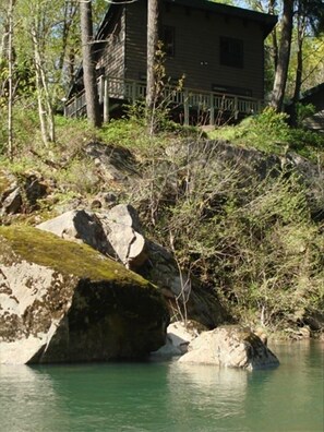 Una baita rustica con una terrazza in legno, circondata da alberi e da una riva rocciosa.