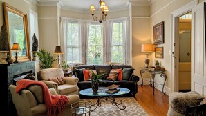Un salon bien éclairé avec une cheminée, une grande fenêtre avec des stores, un lustre et un coin repas avec une table et des chaises.