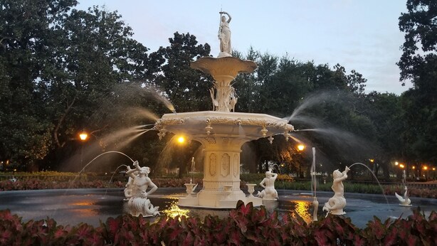 Une fontaine ornée de statues et de jets d’eau, entourée de verdure et illuminée par des lampadaires.