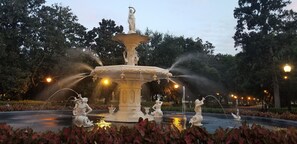 Ein Springbrunnen mit Statuen und Wasserstrahlen, umgeben von Grünflächen und beleuchtet von Straßenlaternen.