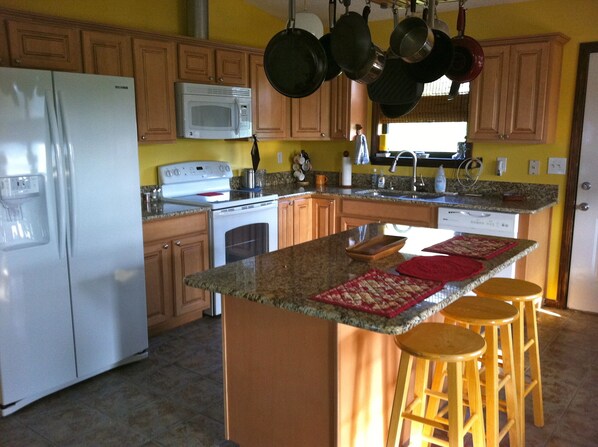 A kitchen with wooden cabinets, a white refrigerator, a stove, and a microwave. There are stools at the island and hanging pots above the stove.