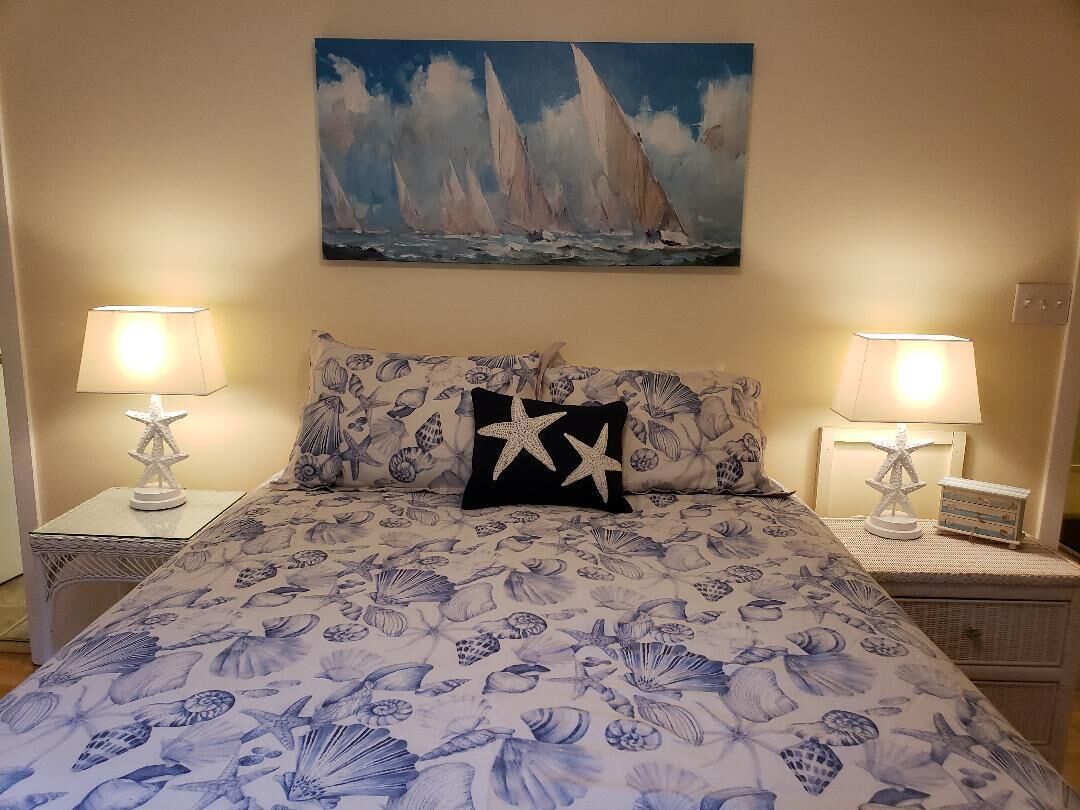 Ein Bett mit Bettdecke mit Muschelmuster, zwei Nachttische mit Lampen und ein Segelboote-Gemälde an der Wand.
