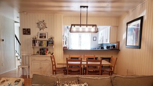 A cozy living room with a brown sofa, wooden dining table, and chairs. There is a kitchen area with white cabinets and a window. - Bethany Beach 3-BR Townhouse With Pool and Tennis - Walking Distance to Beach! (Bethany Beach)