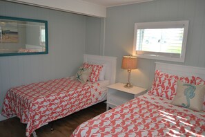 Habitación con dos camas, cada una con una colcha de cuadros rojos y blancos, una mesita de noche con lámpara, un espejo en la pared y una ventana con vistas al exterior.