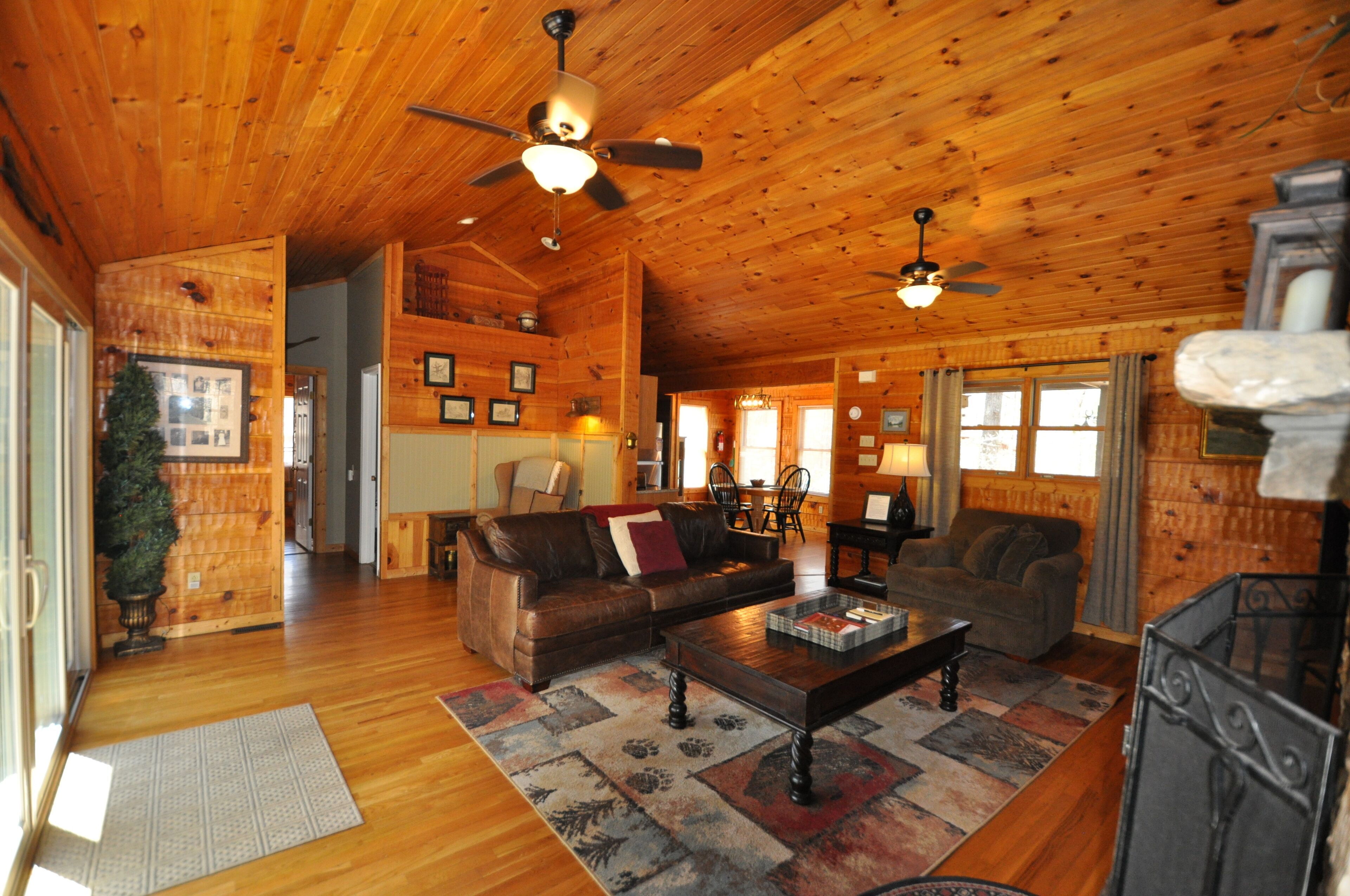 A spacious log cabin interior with a wooden ceiling, two ceiling fans, a living room with a sofa and coffee table, a dining area with a table and chairs, and a kitchen area with a stove.