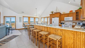 A spacious kitchen with wooden cabinets, a central island with bar stools, and a view of the ocean through large windows. - Oceanfront Dog Friendly Rodanthe Home w/ Pool, Hot Tub, Game Room, Home Theater (Rodanthe)