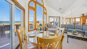 A spacious living room with a large glass table, wooden chairs, and a sofa. There are large windows with wooden frames, offering a view of the beach. - Oceanfront Dog Friendly Rodanthe Home w/ Pool, Hot Tub, Game Room, Home Theater (Rodanthe)
