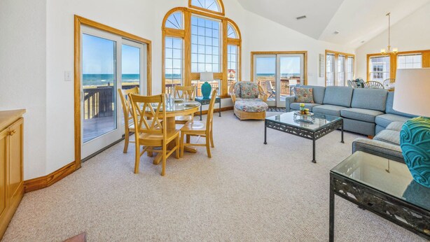 A spacious living room with a large dining table, comfortable seating, and a view of the beach through large windows. - Oceanfront Dog Friendly Rodanthe Home w/ Pool, Hot Tub, Game Room, Home Theater (Rodanthe)