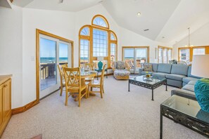 A spacious living room with a large dining table, comfortable seating, and a view of the beach through large windows. - Oceanfront Dog Friendly Rodanthe Home w/ Pool, Hot Tub, Game Room, Home Theater (Rodanthe)
