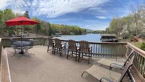 Terraza de madera con mobiliario exterior, una sombrilla roja y vistas a un lago con barcos.