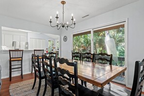 Comedor con mesa y sillas de madera, una lámpara de araña y vista a zonas verdes a través de una puerta corrediza de vidrio.