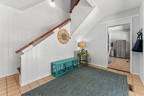 Una escalera con paredes blancas y barandillas de madera, un espejo redondo, un banco color turquesa, una lámpara y una cocina visible a través de un umbral abierto.
