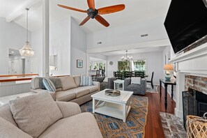 A spacious living room with a ceiling fan, a fireplace, and a television. - Beachwalk Cottage – Steps to Beach, Free Seasonal Pool Heat, Sleeps 10, 4 King Suites (Hilton Head Island)