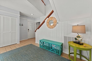 A hallway with a wooden staircase, a round mirror, a teal bench, a yellow table with a lamp, and a door with louvered shutters. - Beachwalk Cottage – Steps to Beach, Free Seasonal Pool Heat, Sleeps 10, 4 King Suites (Hilton Head Island)