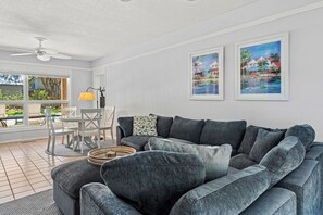 A living room with a grey sectional sofa, a dining table with chairs, and two framed paintings on the wall. - Beachwalk Cottage – Steps to Beach, Free Seasonal Pool Heat, Sleeps 10, 4 King Suites (Hilton Head Island)