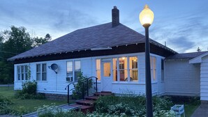 Een wit huis met één verdieping, een donker dak, een veranda en een buitenlamp.