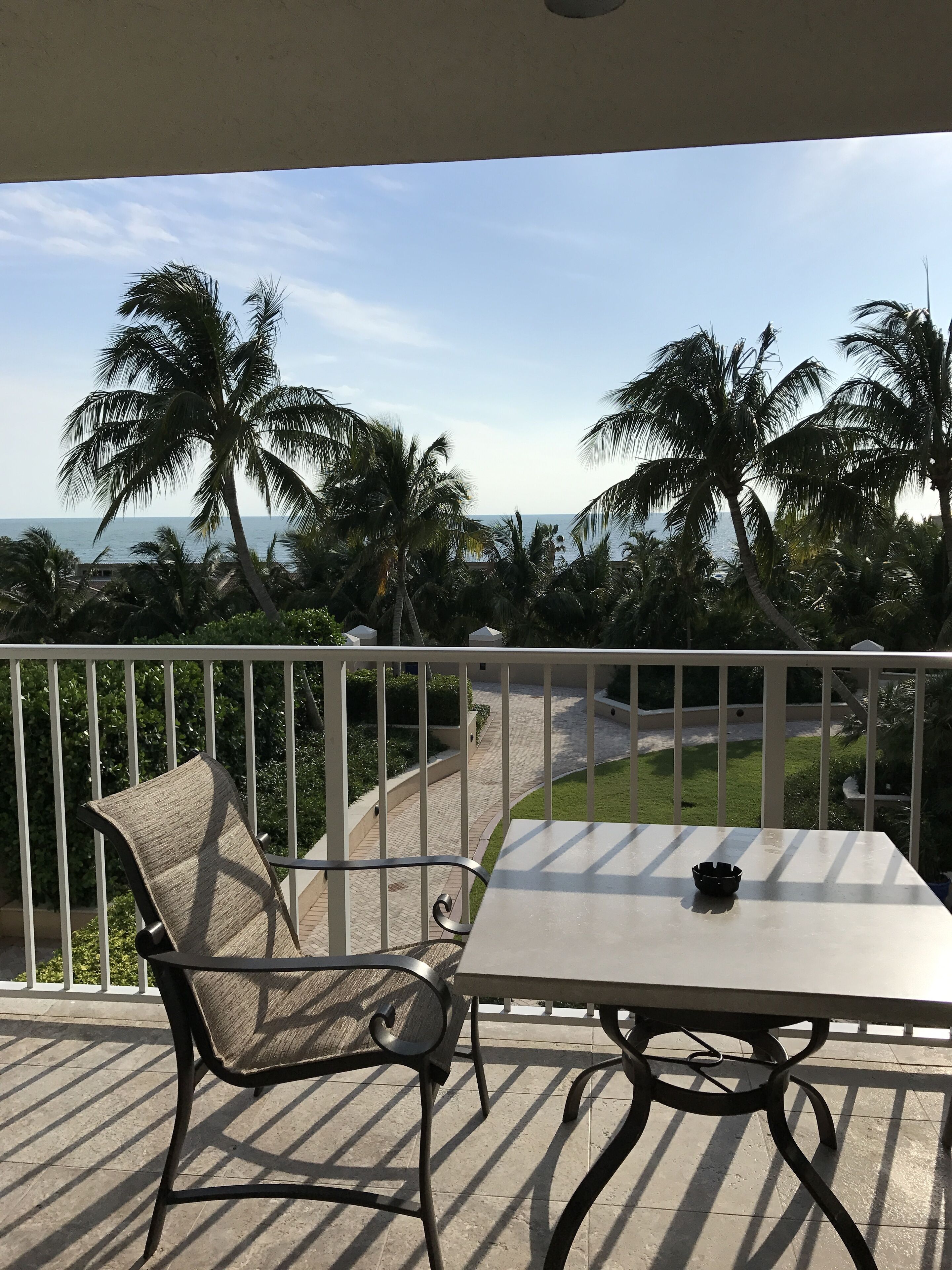 A balcony with a table and chair, overlooking a palm tree-lined path and the sea.