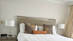 Un lit bien fait, avec du linge de lit blanc, une tête de lit en bois et des coussins décoratifs. Il y a deux tables de chevet avec des lampes et un petit tiroir. La pièce est dotée d’un sol recouvert de moquette et d’un mur blanc uni.