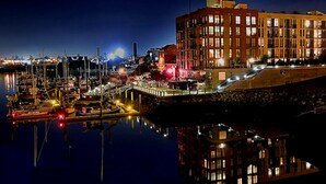 A marina at night with boats docked, a large building with multiple windows, and a calm water surface reflecting the lights. - WATER'S EDGE DOWNTOWN SUNSET WEST BALC, KING BDRM, A/C, 90 DAY MIN, (Victoria)