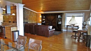 Un amplio espacio habitable con techo de madera, pared de piedra, sofá de cuero, mesa de comedor con sillas y una cocina al fondo.