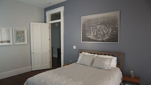 A bedroom with a bed, a nightstand, a framed cityscape picture, and a door leading to another room. - Elegant home steps from St. Charles Ave, parades, and downtown attractions. (New Orleans)