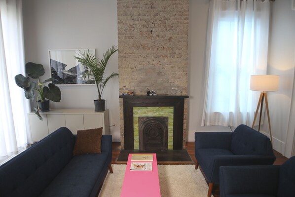 Un salón con dos sofás azul oscuro, una alfombra blanca, una chimenea, un muro de ladrillo, una planta en maceta y una lámpara de pie.