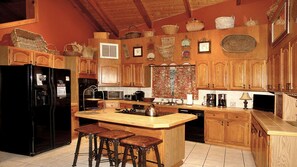 Eine Küche mit Holzschränken, einem schwarzen Kühlschrank, einem Herd und einer Spüle. An der Kochinsel stehen drei Hocker.