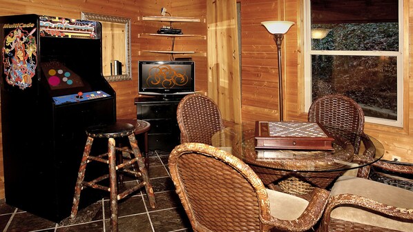 A cozy room with a wooden interior, featuring a gaming console, a television, wicker furniture, and a window with a view of greenery.
