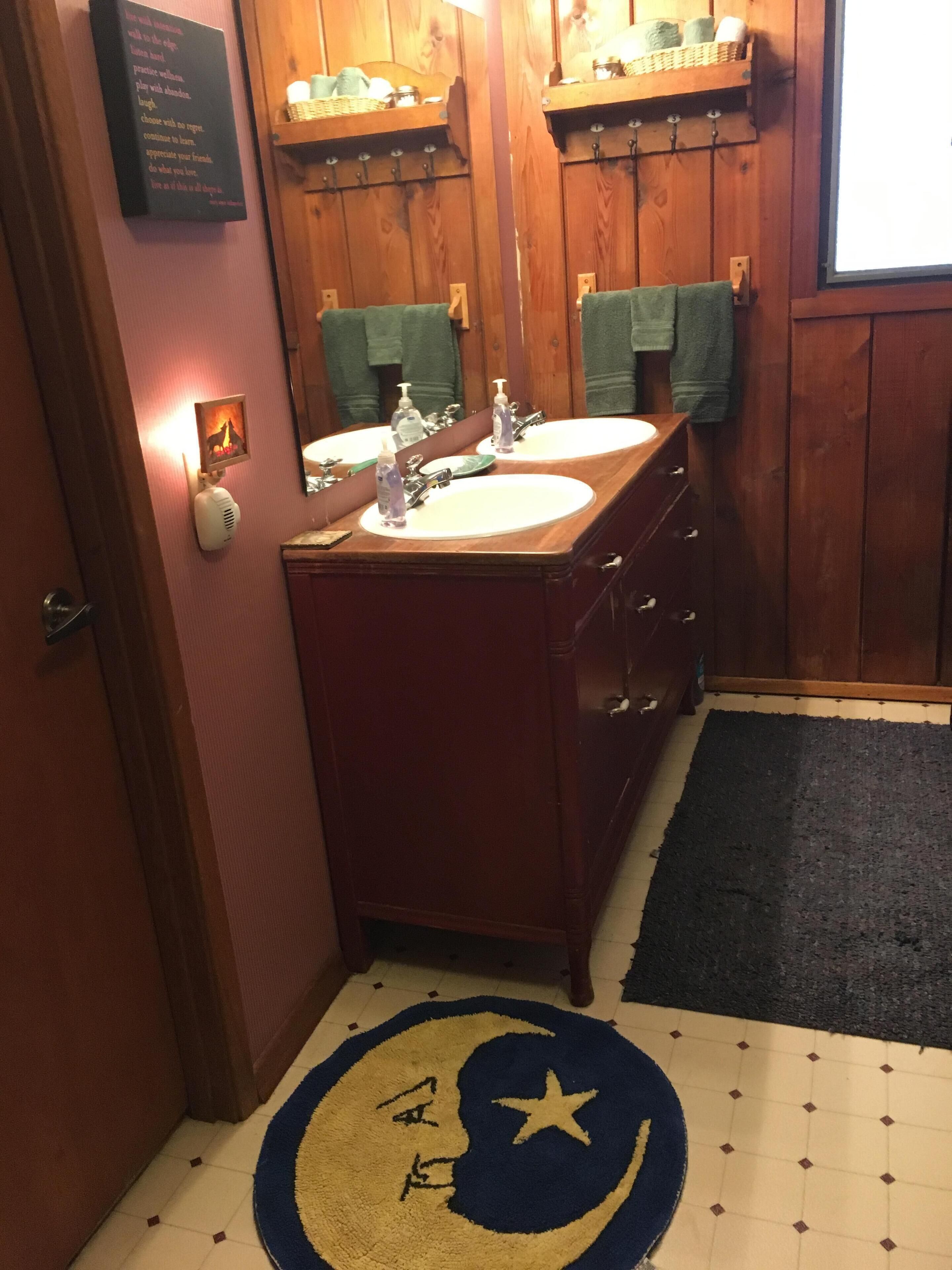Un baño con armarios de madera, dos lavamanos y un espejo. Hay una luz empotrada en la pared y una alfombra con diseño de luna creciente y estrellas en el suelo.