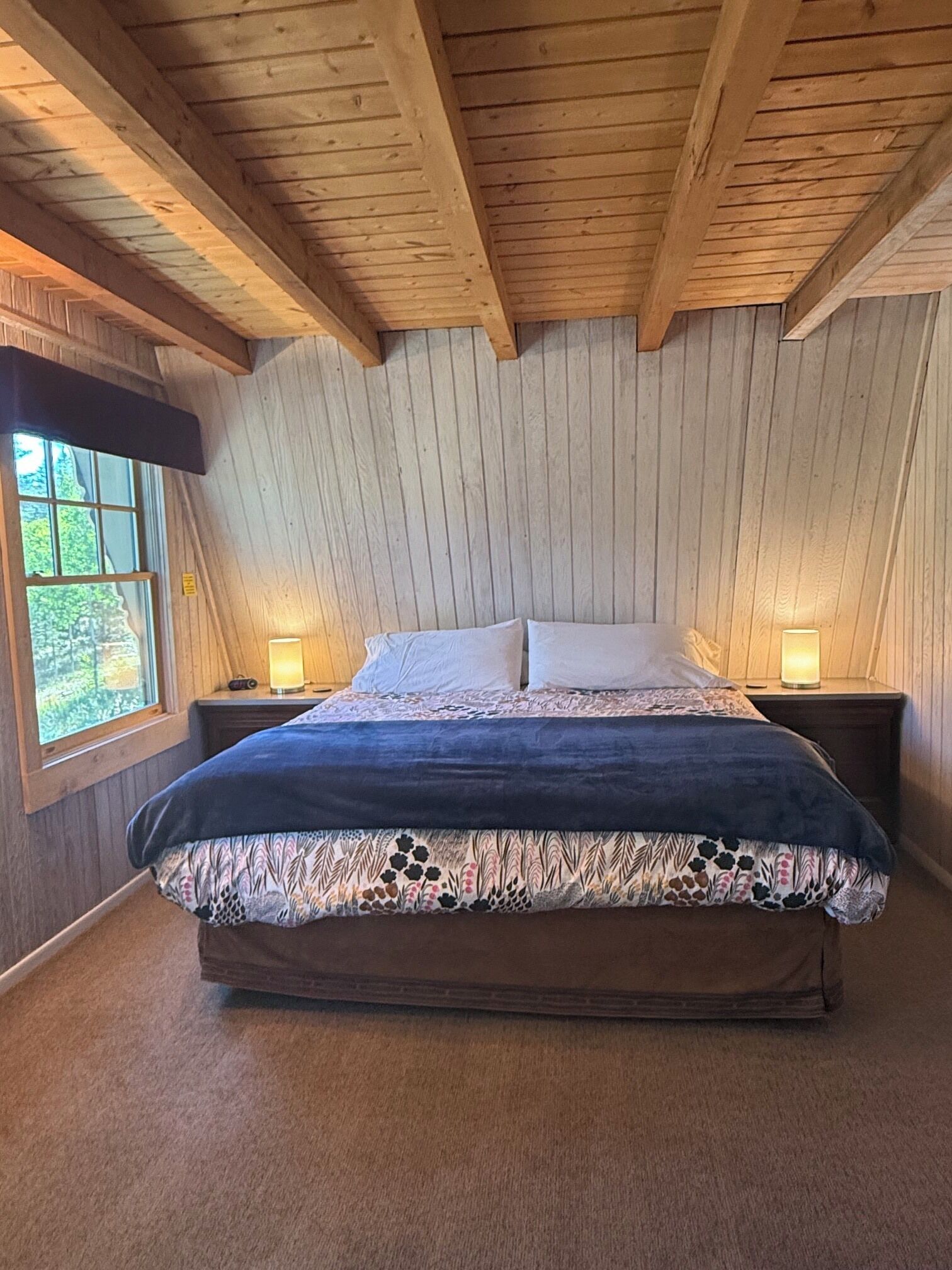 A bedroom with a wooden ceiling, a large bed with a floral bedspread, two bedside lamps, and a window with a view of greenery.