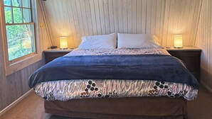A bedroom with a wooden ceiling, a large bed with a floral bedspread, two bedside lamps, and a window with a view of greenery. - Cozy ski chalet 1/2 mile from Main Street, Great Location (Breckenridge)