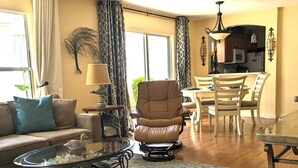 Una sala de estar con un sofá, una mesa de centro de vidrio, un tapete estampado, un sillón de cuero y una zona de comedor con una mesa y sillas.