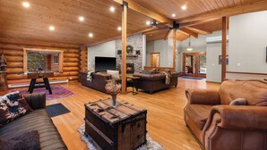 A spacious log cabin interior with a fireplace, leather sofas, wooden floors, and a ceiling fan.
