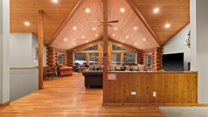 A spacious log cabin interior with a high ceiling, wooden floors, a central seating area with a couch and chairs, and large windows offering views of the outdoors.