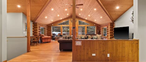 A spacious log cabin interior with a high ceiling, wooden floors, a central seating area with a couch and chairs, and large windows offering views of the outdoors.