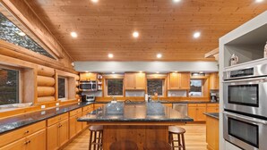 A modern kitchen with wooden cabinetry, a central island with stools, stainless steel appliances, and large windows.
